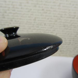May include: Close-up of a black lacquered lid with a small, rounded handle. The lid has a glossy finish with a few small imperfections. The edge of the lid is slightly chipped. A red and black bowl is visible in the background.