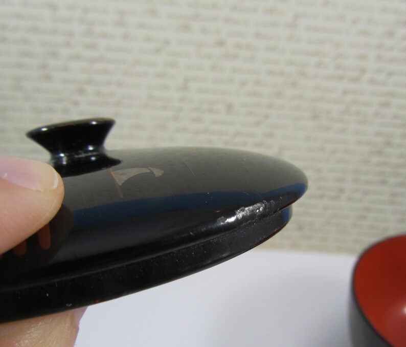 May include: Close-up of a black lacquered lid with a small, rounded handle. The lid has a glossy finish with a few small imperfections. The edge of the lid is slightly chipped. A red and black bowl is visible in the background.