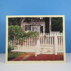 May include: A rectangular greeting card featuring a printed illustration of a white house with a porch, a white picket fence, and a brick walkway. A green vine with orange trumpet-shaped flowers grows over the fence, and a metal chain secures the gate.