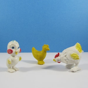 May include: Three small, vintage chicken figurines. One is a white chick with red cheeks and blue eyes. The second is a yellow hen. The third is a white hen with yellow accents and a red comb. All are on a white surface with a blue background.