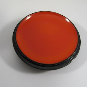 May include: A circular, orange-red object with a glossy surface, contained within a black, circular frame. The item resembles a lid or small dish, with a smooth, reflective appearance. The colour is bright and striking.