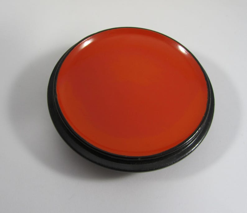 May include: A circular, orange-red object with a glossy surface, contained within a black, circular frame. The item resembles a lid or small dish, with a smooth, reflective appearance. The colour is bright and striking.