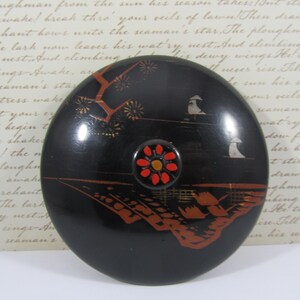 May include: A black, circular decorative lid with a detailed landscape design in red and gold. The lid has a central, flower-shaped knob with red petals and a yellow centre. The design includes trees, buildings, and figures.
