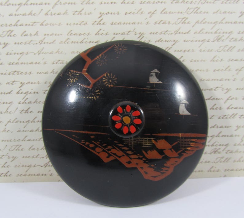 May include: A black, circular decorative lid with a detailed landscape design in red and gold. The lid has a central, flower-shaped knob with red petals and a yellow centre. The design includes trees, buildings, and figures.