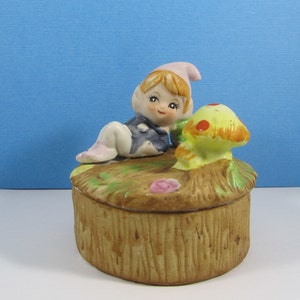 May include: A ceramic trinket box featuring a small elf figurine with a pink hat, lying on a mushroom with red spots. The box is decorated with a brown woodgrain pattern and green leaves.