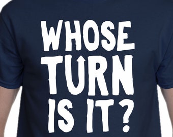 May include: A navy blue short-sleeved t-shirt featuring the white text WHOSE TURN IS IT? printed in a bold, sans-serif font across the chest. The garment has a crew neckline and a standard fit, displaying the text clearly against the dark fabric.
