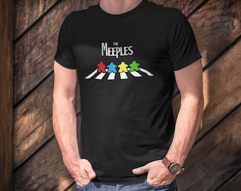 Könnte beinhalten: Schwarzes T-Shirt mit einem Grafikdruck von vier bunten Spielfiguren, die über einen weißen Zebrastreifen laufen. Der Text "THE MEEPLES" befindet sich über der Grafik.