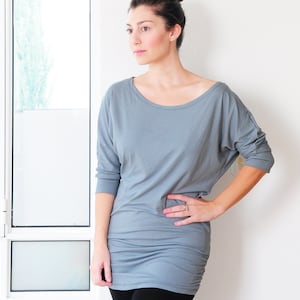 May include: A woman wearing a grey, long-sleeved, loose-fitting shirt with a scoop neck. She is wearing black trousers and has her hair pulled back in a bun.