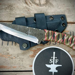 May include: A handmade knife with a camo-patterned handle and a black leather sheath. The knife has a silver blade and a small inscription on the blade. The knife is sitting on a wooden surface.