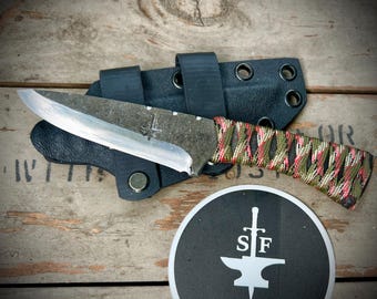 May include: A handmade knife with a camo-patterned handle and a black leather sheath. The knife has a silver blade and a small inscription on the blade. The knife is sitting on a wooden surface.