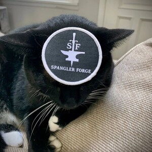 May include: A black and white cat wearing a black and white round patch with the text "SF Spangler Forge" on it.