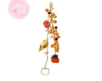 May include: A gold-toned charm bracelet with a variety of decorative elements. It features a red rose, a gold leaf, and a charm of a black cat in an orange pumpkin. The bracelet also includes orange, pink, and black beads.