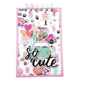 May include: A pink spiral bound notebook with a white background and a colourful pattern of hearts, cupcakes, balloons, and a car with the words "so cute" in black lettering.