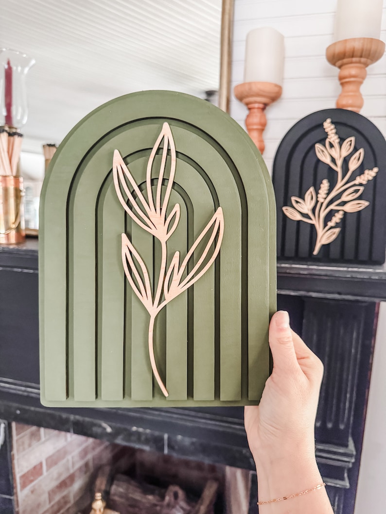Puede incluir: Una pieza de decoraci&oacute;n de pared arqueada verde con un dise&ntilde;o en capas y un acento de hoja marr&oacute;n claro. En el fondo se ve otra pieza de decoraci&oacute;n arqueada negra con un acento floral marr&oacute;n claro. La decoraci&oacute;n la sostiene una persona.