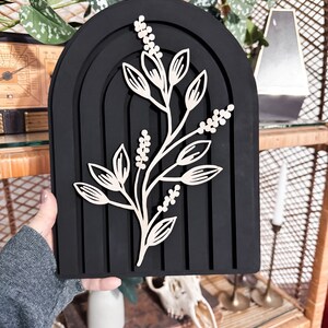 Puede incluir: Decoraci&oacute;n de pared negra en forma de arco con un dise&ntilde;o en capas. Un recorte de estilo bot&aacute;nico de color claro est&aacute; fijado en la parte delantera. La pieza tiene una est&eacute;tica moderna y minimalista, adecuada para la decoraci&oacute;n del hogar.