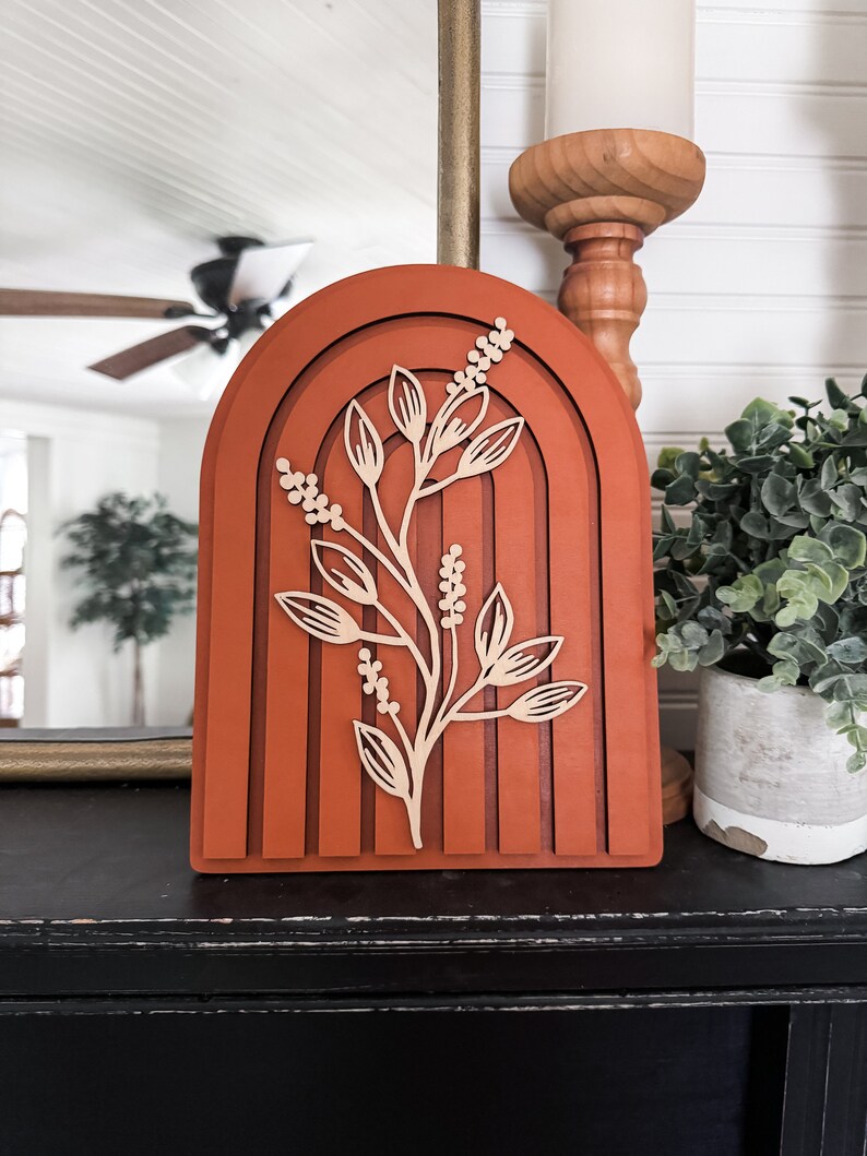 Puede incluir: Una pieza decorativa de madera en capas de color terracota, con un dise&ntilde;o arqueado y un recorte de rama floral. Se coloca sobre una superficie negra, con un candelabro y una planta visibles en el fondo.