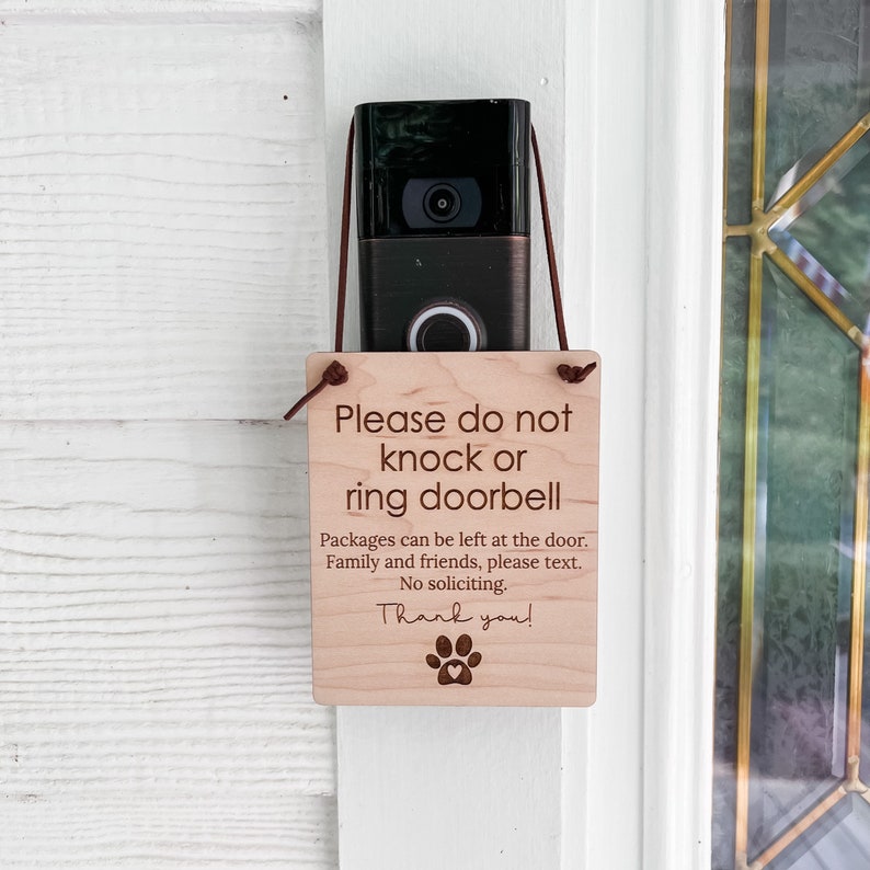 May include: A wooden sign with the text "Please do not knock or ring doorbell" is hanging below a doorbell camera. The sign also includes instructions for package delivery and a paw print. The sign is attached with a brown cord.