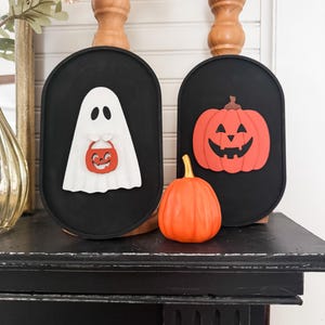 May include: Two black oval-shaped signs with Halloween decorations. The sign on the left has a white ghost holding a red and orange candy bucket. The sign on the right has a red pumpkin with a jack-o-lantern face.