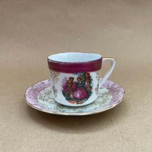 May include: A white teacup and saucer set with a pink and gold floral design. The teacup has a burgundy band around the rim and a painted scene of two people in a garden.
