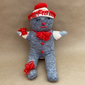 May include: A handmade sock doll with a red and white hat, red yarn for a scarf, and red yarn for arms and legs. The doll is wearing a blue and gray sock for a body.