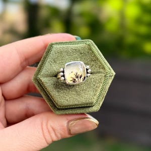 May include: A silver ring featuring a square-shaped dendritic agate gemstone with a translucent, milky white surface and dark, fern-like inclusions. The stone is set in a smooth silver bezel with three small silver spheres on each side.
