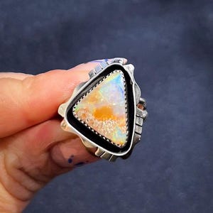 May include: A silver ring featuring a triangular opal stone with a vibrant display of colors, including orange, white, and blue. The opal is set within a black border and surrounded by a detailed silver band.