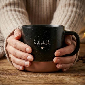 Könnte beinhalten: Eine schwarze, gesprenkelte Kaffeetasse mit braunem Boden, gehalten von Händen. Auf der Tasse steht das Wort "liebedich" und ein Herzsymbol in weißer Schrift. Die Tasse steht auf einer Holzoberfläche.