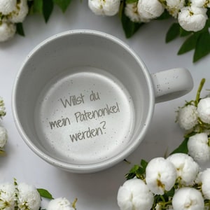 Può includere: Una tazza di ceramica grigia macchiettata con manico. L'interno della tazza presenta il testo tedesco "Willst du mein Patenonkel werden?" in grigio. La tazza è circondata da fiori bianchi e foglie verdi.