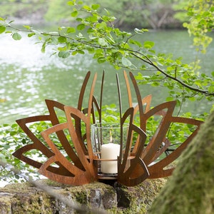 May include: A rusty metal lotus flower candle holder with a glass cylinder and a white candle inside. The candle holder is sitting on a mossy rock near a lake.