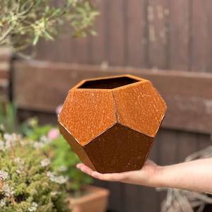 Puede incluir: Un macetero geométrico de metal de color marrón oxidado. El macetero tiene una forma hexagonal con múltiples facetas.