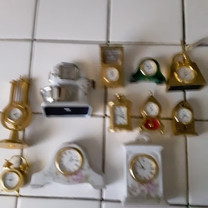 May include: A collection of miniature clocks in various shapes and styles. The clocks feature gold and white tones, with some having floral designs. The collection includes a small mixer and a green clock.