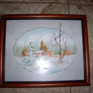 May include: A framed watercolor painting depicting a winter landscape. The artwork features a snow-covered cabin, evergreen trees, and bare trees against a backdrop of a snow-capped mountain and a light blue sky. The frame is a dark brown color.