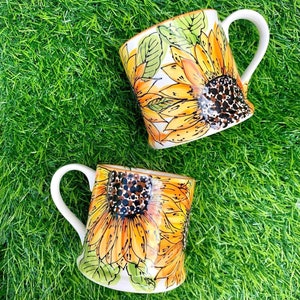 May include: Two white ceramic mugs with sunflower designs. The mugs have white handles and are decorated with yellow and brown sunflowers.