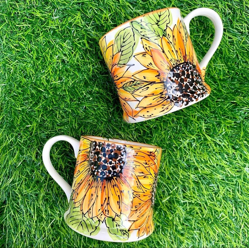 May include: Two white ceramic mugs with sunflower designs. The mugs have white handles and are decorated with yellow and brown sunflowers.