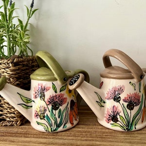 May include: Two ceramic watering cans with floral designs. One is green and white with pink and orange flowers. The other is brown and white with pink flowers.