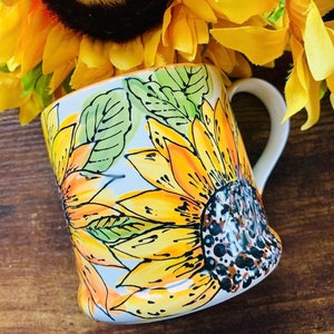 May include: White ceramic mug featuring a hand-painted design of sunflowers in shades of yellow, orange, and green. The mug has a handle and a rounded base.
