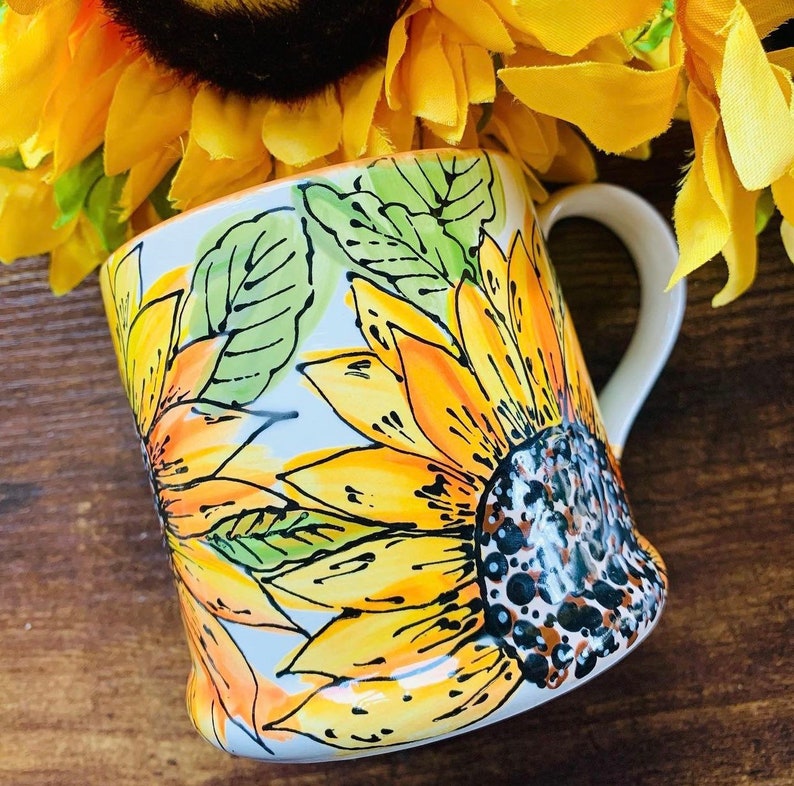 May include: White ceramic mug featuring a hand-painted design of sunflowers in shades of yellow, orange, and green. The mug has a handle and a rounded base.