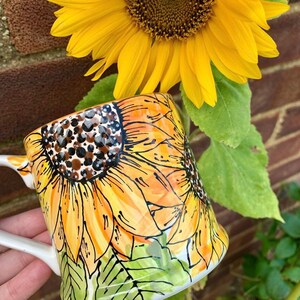 May include: A white ceramic mug featuring a hand-painted design of sunflowers. The sunflowers are yellow, orange and brown with black outlines. The mug has a handle and is resting on a green surface.