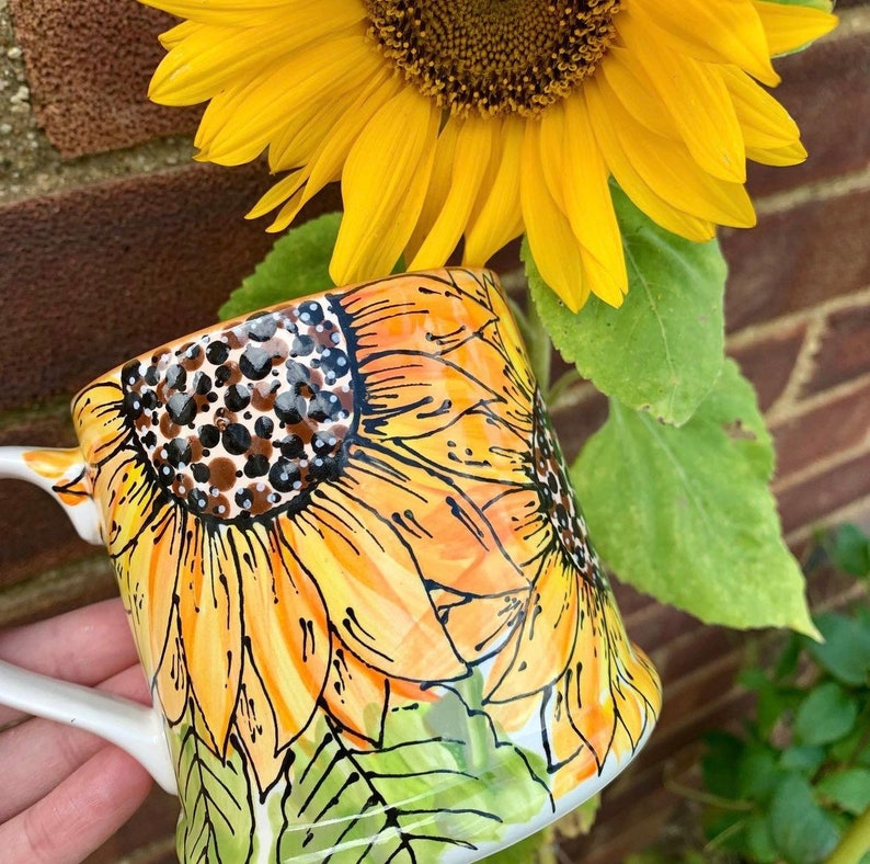 May include: A white ceramic mug featuring a hand-painted design of sunflowers. The sunflowers are yellow, orange and brown with black outlines. The mug has a handle and is resting on a green surface.
