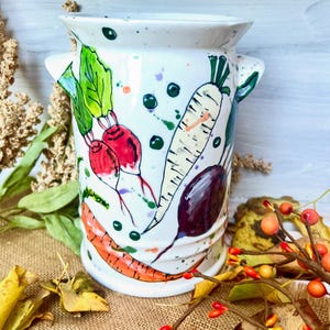May include: White ceramic vase decorated with hand-painted vegetables, such as carrots, radishes, and a parsnip. The vase features a wide opening and two small handles. Green and purple paint splatters add a whimsical touch.