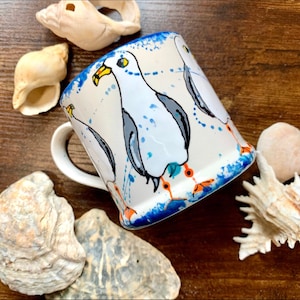 May include: A white ceramic mug with a blue rim and a blue and white splatter design. The mug features a hand-painted design of three seagulls with orange feet and black eyes.