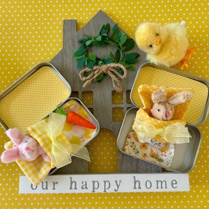 May include: Two small tin boxes with yellow polka dot fabric lining, each containing a stuffed animal. One box has a pink bunny with a carrot, the other has a brown bunny. A yellow chick sits on top of a wooden house with a green wreath. The sign below reads "our happy home".