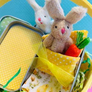 May include: A yellow and white polka dot fabric bed with a yellow ribbon bow, a brown bunny with a carrot, and a white bunny peeking out of the bed. The bed is in a metal tin with a green and yellow polka dot lid. The tin is sitting on green and yellow shredded paper.