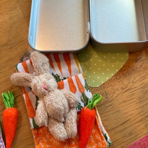 May include: A small, plush brown bunny rabbit laying on a fabric pouch with a carrot print. The pouch is open and there are two small, orange carrots next to it.