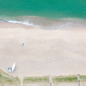 May include: An aerial view of a sandy beach with turquoise water and white waves. A small white boat is on the sand, and a person is lying on a towel.