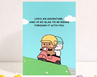 May include: A greeting card with a light blue background and a cartoon illustration. The image features two characters riding a pink scooter, with the text "LIFE'S AN ADVENTURE, AND I'M SO GLAD TO BE RIDING THROUGH IT WITH YOU."