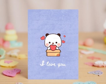 Könnte beinhalten: Eine hellblaue Karte mit einem Comic-Panda, der ein rotes Herz in einer Schachtel hält. Der Text "Ich liebe dich" ist in weißer Kursivschrift auf der Karte geschrieben.