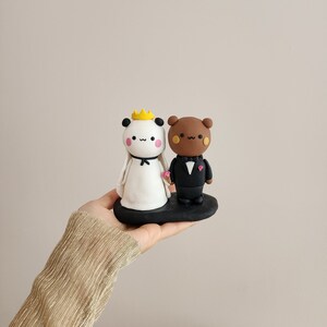 May include: A white panda and a brown bear figurine wearing wedding attire. The panda is wearing a white dress and a gold crown. The bear is wearing a black tuxedo. The figurines are standing on a black base.