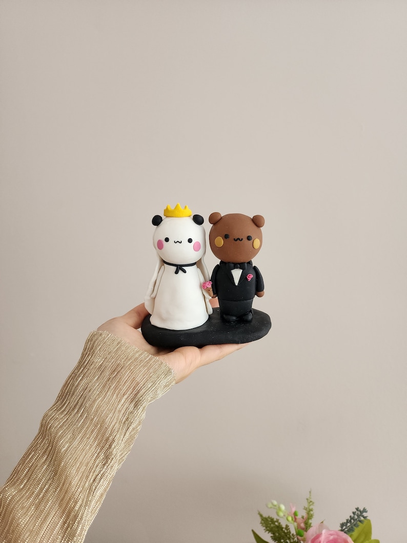 May include: A white panda and a brown bear figurine wearing wedding attire. The panda is wearing a white dress and a gold crown. The bear is wearing a black tuxedo. The figurines are standing on a black base.