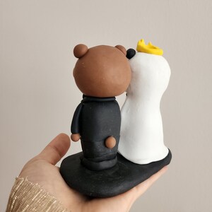May include: A brown bear wearing a black suit and a white polar bear wearing a white dress and a yellow crown. The bears are standing on a black base.
